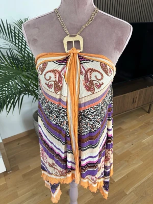Mönstrad strandklänning i orange och lila Patrizia pepe - Säljer en färgglad strandklänning i boho-stil med orange, lila och vita toner. Klänningen är axelbandslös med knyt fram och har ett mixat mönster av ränder och paisley. Perfekt för sommardagar och semester.