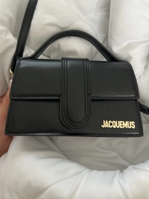 Jacquemus väska  - Säljer en svart Le Chiquito handväska från Jacquemus i slätt läder med guldfärgad logga framtill. Väskan har ett topphandtag, avtagbar axelrem och stilren, boxig form. Perfekt för dig som gillar trendiga accessoarer och minimalistisk design.