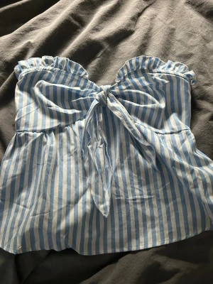 Blåvit randig bandeau topp med knyt - Säljer en söt bandeau topp i blåvitt randigt med volangkant upptill och knyt framtill. Toppen är ärmlös och har en luftig passform, perfekt för sommaren!💓 (Oanvänd)