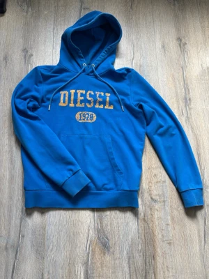 Blå hoodie från Diesel med tryck - Snygg blå hoodie från Diesel med gul text och årtalet 1978 tryckt på bröstet. Klassisk känguruficka framtill, ribbade muddar och justerbar huva med dragsko. Perfekt för en avslappnad och cool stil.