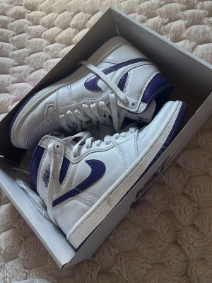 Nike Air Jordan 1 High vit/lila - Säljer ett par Nike Air Jordan 1 i vitt och lila. Storlek 37,5 (men skulle säga något små i storleken). Skorna är använda ett fåtal gånger, och kommer med kartongen de köptes i. 