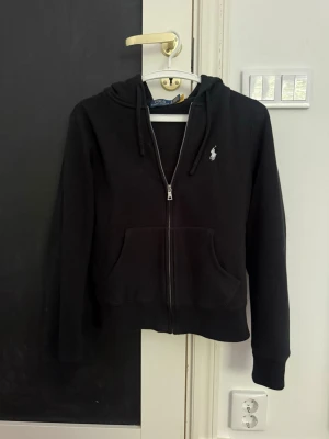 Svart zip-hoodie Ralph lauren - Snygg svart hoodie med dragkedja och huva. Tröjan har två fickor framtill och en liten vit logga broderad på bröstet. Perfekt för en avslappnad stil och enkel att matcha med jeans eller mjukisbyxor.