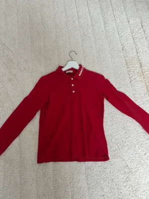 Röd Moncler långärmad polotopp - Säljer en röd långärmad polotopp från Moncler med vit logga på kragen och tre silverfärgade knappar framtill. Klassisk pikékrage och stilren design, perfekt för dig som vill ha något snyggt och bekvämt. Ny pris ca 2000kr. Passar mig inte längre. Storlek 14A och passar storlek S