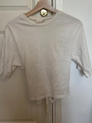 Vit topp med knytband bak - Säljer en enkel och stilren vit topp med korta ärmar och rund hals. Toppen har snygga knytband i ryggen som ger en extra detalj. Perfekt att matcha med jeans eller kjol för en clean look.