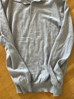 Grå långärmad Piké John Henric - Snygg grå långärmad tröja från John Henric med krage och knappar vid halsen. Tillverkad i mjukt material och har ribbade muddar vid ärmslut och nederkant. Perfekt för en stilren och avslappnad look. Sitter mer som xs