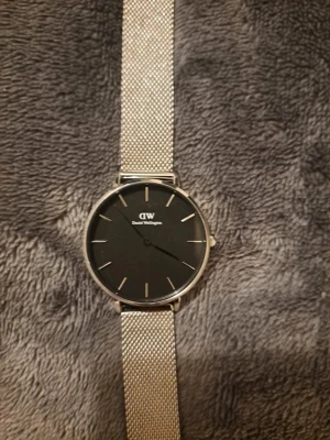 Silverfärgad klocka från Daniel Wellington - Stilren klocka från Daniel Wellington med svart urtavla och silverfärgat mesharmband. Minimalistisk design med tunna silvermarkeringar och DW-logga på urtavlan. Perfekt accessoar för en clean look.
