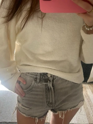 Grå denimshorts med fransig kant - Säljer ett par grå denimshorts med rå, fransig kant och slitna detaljer. Shortsen har hög midja, klassiska fem fickor och knappgylf. Perfekta för varma dagar och ger en avslappnad vibe till din outfit.