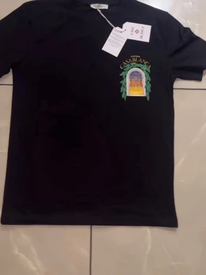 Svart t-shirt med tryck från Casablanca - Snygg svart t-shirt från Casablanca med färgglatt tryck på bröstet som föreställer en byggnad och gröna blad. T-shirten har klassisk passform och rund hals. Perfekt för dig som vill ha en unik och trendig look.