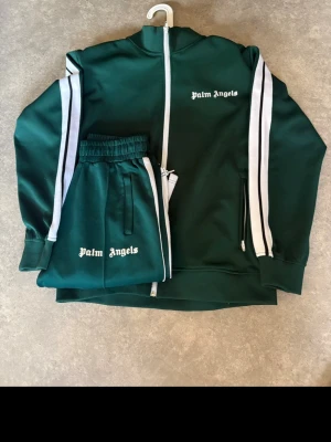 Grönt loungewear-set Palm Angels - Mörkgrönt loungewear-set från Palm Angels med vita ränder längs ärmar och byxben. Setet består av en ziptröja med hög krage och matchande byxor med resår i midjan. Båda delarna har Palm Angels-logga broderad i vitt. Perfekt för chill dagar hemma.