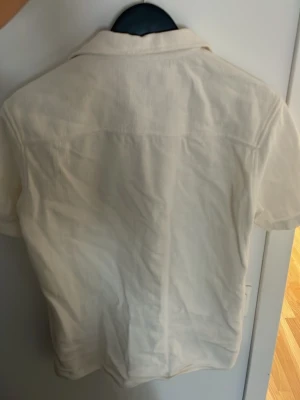 Vit kortärmad skjorta från Zara - Säljer en stilren vit kortärmad skjorta från Zara. Skjortan har klassisk krage, knäppning framtill och är tillverkad i ett lätt, strukturerat tyg som känns luftigt. Perfekt för varma dagar och enkel att matcha med olika outfits.