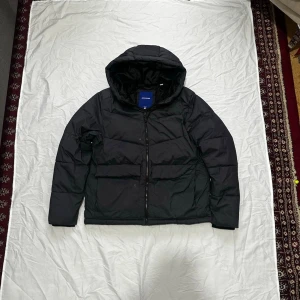 Jack & Jones jacka  - Snygg svart pufferjacka med stor huva och dragkedja framtill. Jackan har två stora fickor på sidorna och en boxig passform som ger en modern look. Perfekt för kalla vinterdagar och riktigt varm tack vare det tjocka materialet.