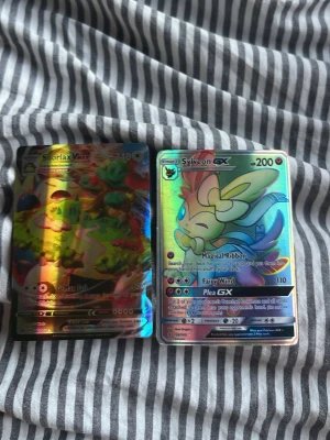 Pokémonkort Snorlax VMAX & Sylveon GX - Två holografiska Pokémonkort: Snorlax VMAX med färgglad bakgrund och Sylveon GX i pastellfärger. Båda korten har glansig yta och detaljerade illustrationer. Perfekt för samlare eller Pokémonfans.