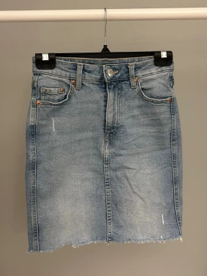 Ljusblå jeanskjol med rå kant - Säljer en ljusblå jeanskjol i klassisk modell med fem fickor, bälteshällor och knappstängning fram. Kjolen har en rå, fransig kant nedtill och subtila slitningar för en avslappnad look. Perfekt till sneakers eller boots.
