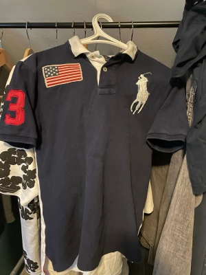 Marinblå pikétröja från Ralph Lauren - Snygg marinblå pikétröja från Polo Ralph Lauren med vit krage, stor logga på bröstet, amerikansk flagga på höger sida och röd siffra 3 på ärmen. Klassisk passform och mjukt bomullsmaterial. Perfekt för en sportig och avslappnad look.