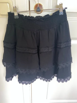 Svart kortkjol från Ginatricot i storlek Small - Supersöt svart kortkjol med bred resår i midjan och flera lager volanger dekorerade med spetsdetaljer. Kjolen har en luftig och flowy känsla, perfekt för att styla med en enkel topp eller hoodie.