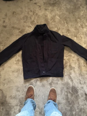 Mörkblå overshirt med fickor - Snygg mörkblå overshirt med hög krage och fyra stora fickor framtill. Rak passform och knäppning med knappar. Perfekt att slänga över en t-shirt för en avslappnad look. Materialet ser ut att vara bomull eller en bomullsblandning.