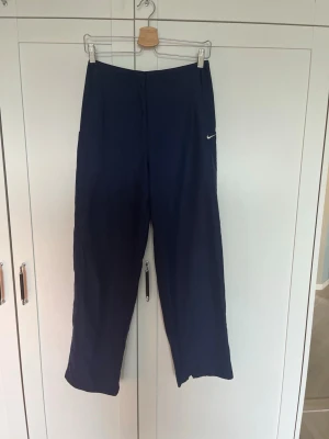 Vintage Nike Träningsbyxor - Snygga mörkblå vintage träningsbyxor från Nike med rak passform och diskret vit logga broderad vid fickan. Byxorna har en enkel design med sidofickor och är tillverkade i ett mjukt och bekvämt material som passar perfekt för chill dagar och gym. Byxorna är unisex så passar XS för killar och S för tjejer