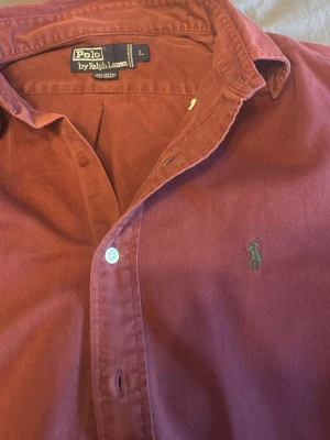 Vinröd skjorta från Polo Ralph Lauren - Säljer en vinröd skjorta från Polo Ralph Lauren med klassisk krage och knäppning framtill. Skjortan har lång ärm och den ikoniska broderade loggan på bröstet. Perfekt för dig som gillar stilrena plagg med en tidlös touch.
