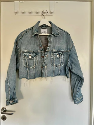 Cropped jeansjacka från ZARA - Cropped ljusblå jeansjacka från ZARA med rå kant nertill och två bröstfickor med knapp. Klassisk krage och knappstängning framtill. Jackan har en avslappnad passform och detaljer i silverfärgade knappar. Perfekt för en trendig och avslappnad look.
