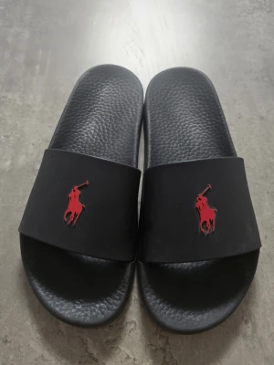 Svarta slides med röd logga - Säljer ett par svarta slides från Polo Ralph Lauren med den klassiska röda hästloggan på ovandelen. Sandalerna har en bred rem och en strukturerad fotbädd i syntetmaterial för extra komfort. Perfekta för chill dagar eller till stranden.