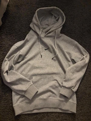 Grå hoodie från Nike med huva - Säljer en stilren grå hoodie från Nike med klassisk huva och dragsnören. Hoodien har en liten svart Nike-logga på bröstet och en stor magficka framtill. Tillverkad i mjukt bomullsmaterial och har ribbade muddar vid ärmar och nederkant.
