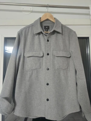 Grå overshirt från H&M - Stilren grå overshirt från H&M i mjukt material. Klassisk rak passform med två stora bröstfickor med lock och svarta knappar framtill. Perfekt lager-på-lager-plagg med krage och långa ärmar.