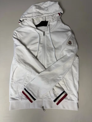 Vit hoodie från Moncler med dragkedja - Snygg vit hoodie från Moncler med hel dragkedja framtill och huva. Ärmsluten och huvan har ränder i rött, vitt och blått. Moncler-logga på vänster ärm. Tillverkad i mjukt bomullsmaterial och har två fickor framtill.   ⚠️den är äkta! ⚠️