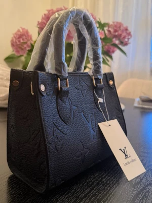 Svart LV väska - Snygg svart handväska från Louis Vuitton med präglat monogrammönster och guldfärgade detaljer. Väskan har två handtag och en kompakt, boxig form. Tillverkad i strukturerat läder och levereras med tagg. Perfekt accessoar för en trendig look. Skriv till mig om du är intresserad för fler bilder. Aldrig använd så nyskick. Not real