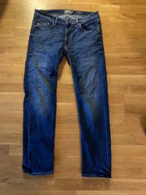 Mörkblå regular fit dressmann jeans - Säljer ett par klassiska mörkblå jeans med rak passform och fem fickor. Jeansen har normal midja och subtila slitningar för en avslappnad look. Tillverkade i denim med kontrastsömmar och knappgylf.