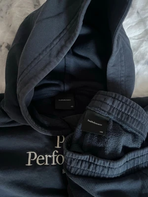 Mörkblått loungewear-set Peak Performance - Snyggt mörkblått loungewear-set från Peak Performance med matchande hoodie och byxor. Båda plaggen har broderad logga framtill och mjuk insida. Hoodien har huva och byxorna har resår i midjan och vid benslut. Perfekt för chill dagar hemma.