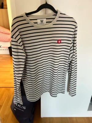 Lång armad tröja by Wood Wood  - Snygg långärmad tröja från double A med svartvita horisontella ränder och rund halsringning. Tröjan har en liten röd patch med ett vitt W på bröstet. Perfekt för en avslappnad och trendig stil.