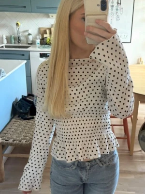 Vit prickig blus med lång ärm - Snygg vit blus med svarta prickar och långa ärmar. Blusen har smock i midjan som ger en figurnära passform och volang nedtill. 