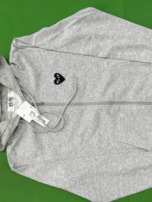 Grå hoodie från Comme des Garçons Play - Snygg grå hoodie från Comme des Garçons Play med dragkedja och svart hjärta med ögon broderat på bröstet. Tillverkad i mjuk bomullsblandning och har justerbar huva med snören samt två fickor framtill. Perfekt för en chill och trendig look.
