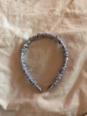 Ljusblått satin-diadem - Ett snyggt diadem i ljusblått satin med rynkad design. Perfekt accessoar för att lyfta din look med en trendig touch. Diademet har en mjuk glans och är täckt av ett slätt tyg som ger en lyxig känsla. Något skadad längs ner, dock syns det inte bakom örat.