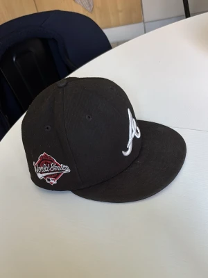 Svart new era  Atlanta Braves keps  - Säljer en svart keps från New Era med Atlanta Braves-logga broderad i vitt framtill och World Series-märke på sidan. Klassisk flat brim och strukturerad form, perfekt för dig som gillar sportig streetstyle. Materialet är bomullstyg.