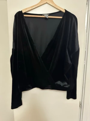 Svart omlottopp i sammet från Lindex - Snygg svart omlottopp i mjuk sammet från Lindex. Toppen har lång ärm och djup v-ringning framtill, perfekt för att lyfta din outfit med en elegant touch. Passformen är lite loose och materialet känns lyxigt mot huden.