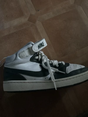 Nike vita och svarta midsneakers - Nike sneakers i vitt och svart med klassisk swoosh på sidan. Skon har en mellanhög siluett med kardborreband upptill och vita snören. Ovandelen är i läder och mesh för extra komfort och stil. Perfekt för dig som gillar streetwear och sportig look. (Pris kan disskuteras)