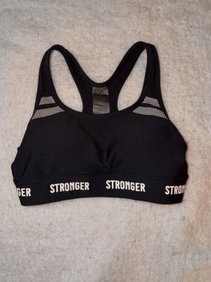 Svart sport-bh från Stronger XS - Snygg svart sport-bh från Stronger i storlek XS. Bh:n har brottarrygg, vita detaljer och logga runt resåren. Materialet är stretchigt och består av 75% nylon och 25% elastan, perfekt för träning och aktivitet.