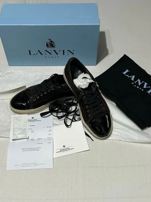 Svarta sneakers från Lanvin - Snygga svarta sneakers från Lanvin med lackade tåpartier och ovandel i mocka. Skorna har svarta skosnören, vit sula och klassisk låg profil. Kommer med originalkartong och dustbag. Perfekta för dig som gillar exklusiva detaljer och stilren design.