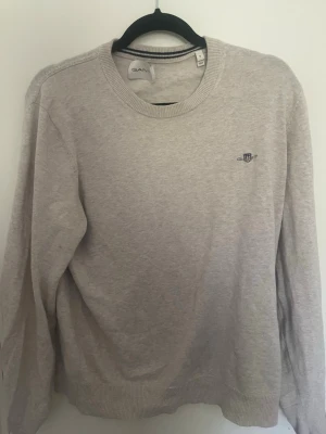 Beige stickad tröja från GANT - Snygg beige stickad tröja från GANT med rund halsringning och diskret broderad logga på bröstet. Tröjan har ribbade muddar vid ärmslut och nederkant, perfekt för en stilren och avslappnad look. Hål under armhålan som kan sprättas upp. Nypris ca 1000 kr