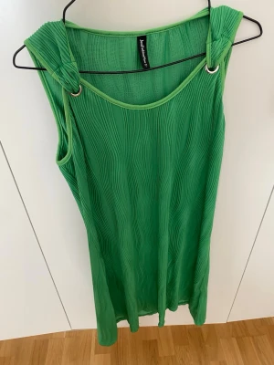 Grön ärmlös kortklänning - Snygg grön kortklänning med vågigt plisserat tyg och ringdetaljer vid axlarna. Klänningen är ärmlös och har en avslappnad passform som ger en cool och modern look. Perfekt för varma dagar och enkel att styla med accessoarer. Står stl L men passar mer S/m om man vill ha den som på bilden lösare 
