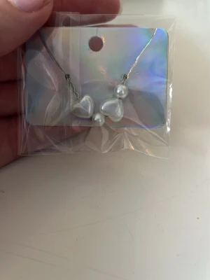 Sött halsband med pärlor och hjärtan - Ett gulligt halsband med vita pärlor och två hjärtformade detaljer. Kedjan är tunn och silverfärgad, vilket ger ett stilrent intryck. Perfekt accessoar för att lyfta din outfit med en söt touch.
