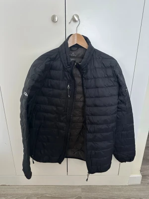 Svart pufferjacka från Elevate - Snygg svart pufferjacka från Elevate med quiltad design och hög krage. Jackan har dragkedja framtill, två sidofickor med dragkedja och diskret logga på ärmen. Perfekt för kalla vinterdagar och riktigt lätt att bära.