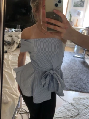 Blåvit off shoulder topp med knytband - Säljer en blåvit randig off shoulder topp med bred volang upptill och knytband i midjan. Toppen har en peplumformad nederdel som ger en snygg siluett. Perfekt för dig som gillar trendiga detaljer och en feminin look.