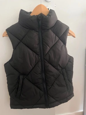 Svart quiltad dunväst med hög krage - Stilren svart dunväst med hög krage och dragkedja framtill. Västen har ett quiltat mönster i stora rutor och två sidofickor. Perfekt att slänga över en hoodie eller tröja för extra värme. Materialet är mjukt och puffigt för en skön känsla.