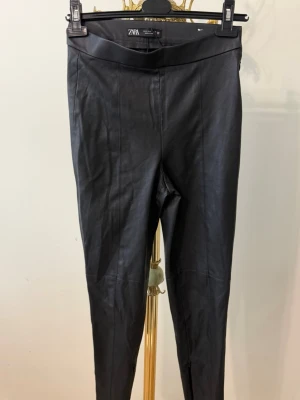 Svarta leggings i fuskläder från ZARA - Snygga svarta leggings i fuskläder från ZARA med hög midja och dragkedja vid benslut. Tight passform och stilren design som ger en edgy look. Perfekta att matcha med oversized tröja eller crop top.