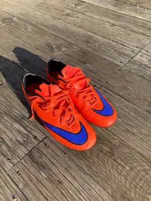 Oranga Nike mercurial fotbollsskor med blå detalj - Säljer ett par snygga fotbollsskor från Nike i starkt röd färg med stora blå swoosh-loggor på tån. Skorna har klassisk snörning och låg profil, samt svart insida. Perfekta för dig som vill sticka ut på planen!