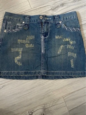Kort jeanskjol med broderade detaljer - Säljer en kort jeanskjol i blå denim med broderade och sydda detaljer i kontrastfärg. Kjolen har fickor både fram och bak, bälteshällor och en knapp framtill. Snygga slitningar och dekorativa sömmar ger kjolen en unik och trendig look.