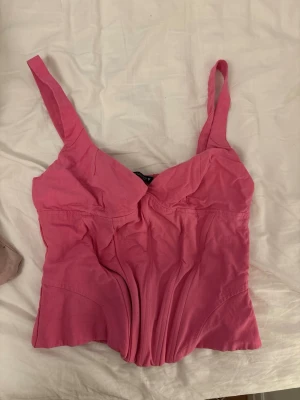 Rosa korsettopp med breda axelband - Snygg rosa korsettopp med breda axelband och markerade sömmar framtill som ger en cool siluett. Toppen har smockad rygg för extra komfort och sitter tajt. Perfekt att styla med jeans eller kjol för en trendig look.