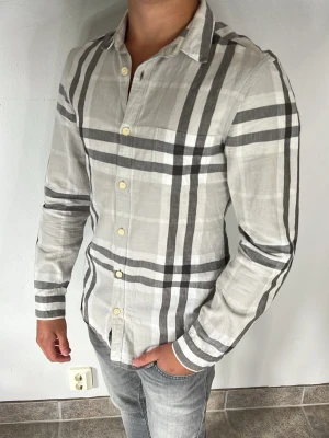 Burberry skjorta - Snygg ljusgrå skjorta med stora mörkgrå rutor och klassisk krage. Skjortan har långa ärmar och stängs med ljusa knappar framtill. Perfekt för en stilren och modern look.